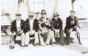 In the foreground there are five older white men sitting on a bench. In the background in front of a building. The photo is black and white.